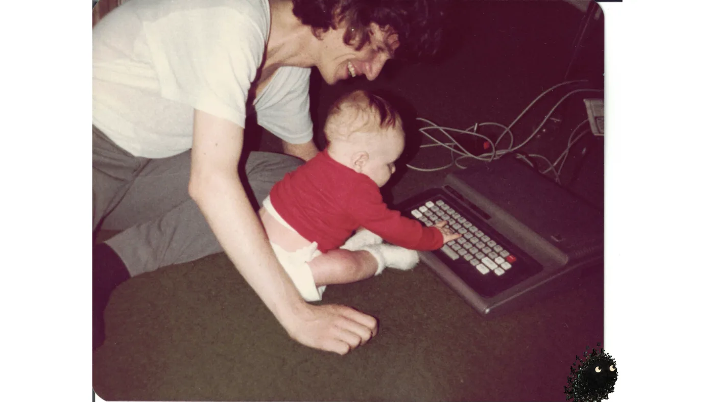 picture: an early-eighties snapshot — a laughing young man kneeling on brown carpet, holding up a baby in red who reaches for the keyboard of a TRS-80 Color Computer | picture: Sili, in the bottom right, moving right